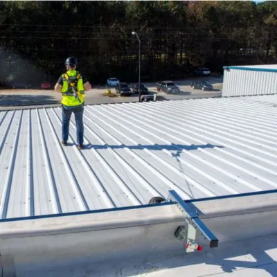 Worker wearing a safety harness and standing on a commercial metal roof with an anchor tie-down system for fall protection while performing minor roof maintenance